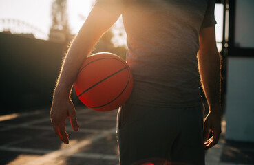 Wall Mural - Male holds a basketball at their side, bathed in warm sunlight, with the focus on the ball and their arm. The soft, natural lighting creates a calm and contemplative atmosphere on the outdoor court