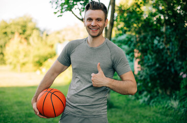 Wall Mural - A smiling man in a gray t-shirt holds a basketball in one hand while giving a thumbs-up with the other. The capturing of a positive and energetic moment, is perfect for sports or lifestyle themes.