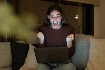 Excited young woman with a surprised expression looks at her laptop screen while sitting alone on a sofa at night. She is thrilled and happy, browsing the internet at home, celebrating  achievement