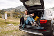 © maxbelchenko - A joyful young woman sitting by the open trunk of her car in a mountainous landscape during a sunny day, enjoying the great outdoors. Journey by car.