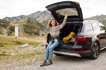 Wall Mural - A joyful young woman sitting by the open trunk of her car in a mountainous landscape during a sunny day, enjoying the great outdoors. Journey by car. 