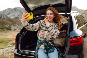 Wall Mural - Young traveler woman taking a selfie by her car in the mountains during sunset, showcasing her stylish outfit and joyful spirit. Lifestyle, travel, tourism, nature, active life.