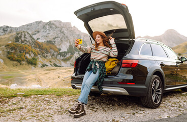 Young traveler woman taking a selfie by her car in the mountains during sunset, showcasing her stylish outfit and joyful spirit. Lifestyle, travel, tourism, nature, active life.