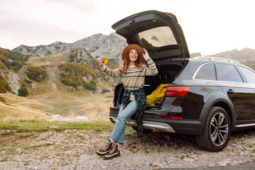 Wall Mural - Young traveler woman taking a selfie by her car in the mountains during sunset, showcasing her stylish outfit and joyful spirit. Lifestyle, travel, tourism, nature, active life.