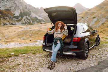 Wall Mural - Young traveler woman taking a selfie by her car in the mountains during sunset, showcasing her stylish outfit and joyful spirit. Lifestyle, travel, tourism, nature, active life.