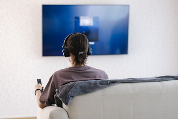 Back view of teenage boy puts on wireless headphones and connect them to tv for watching show. Big screen 