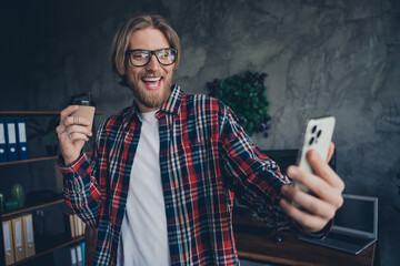 Photo of young successful businessman wearing checkered shirt holding coffee to go photographing his day at office indoors