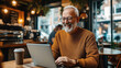 © altitudevisual - Elderly man with glasses and a beard smiling and working on a laptop in a cozy coffee shop, with a takeaway coffee cup on the table.