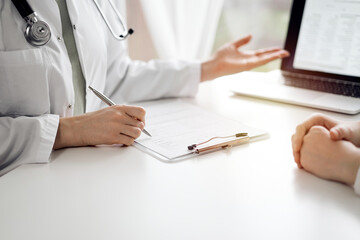 Wall Mural - Doctor and patient sitting and discussing something near each other at the white desk in clinic. Female physician is pointing into laptop screen. Medicine concept