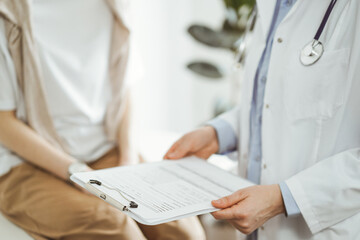 Wall Mural - Doctor and patient discussing perfect health exam results. Friendly physician keeping with hands clipboard with medical papers near young woman. Medicine concept