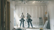 © Gita - Two construction workers in safety gear preparing and finishing drywall installation in a room under renovation. Ideal for themes related to construction, remodeling, and interior design.