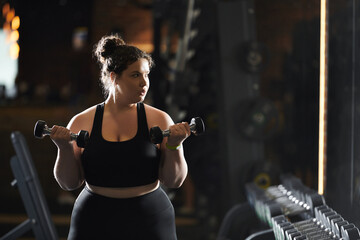 Wall Mural - A plus-size woman confidently lifts dumbbells, celebrating strength and wellness in an energetic gym atmosphere.