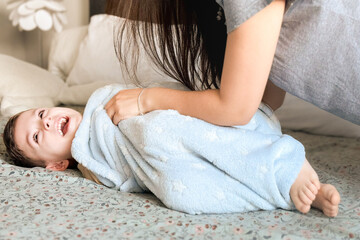 Wall Mural - Mom tickles the feet of her little son, wrapped in a blue blanket. Mom and child have fun together in a cozy bedroom at home. Happy children with mother. Bright photos of the baby's childhood