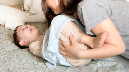 Wall Mural - Mom tickles the feet of her little son, wrapped in a blue blanket. Mom and child have fun together in a cozy bedroom at home. Happy children with mother. Bright photos of the baby's childhood