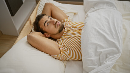 Canvas Print - A thoughtful young hispanic man with a beard lies in bed, gazing upward in a cozy bedroom setting.