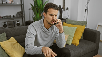 A concerned young hispanic man with a beard in casual attire making a phone call from a cozy living room couch.