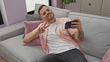 Wall Mural - A handsome young man with a beard takes a selfie while making a peace sign and relaxing on a grey sofa indoors.