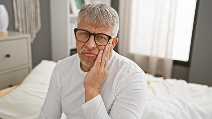 Canvas Print - A pensive, grey-haired man wearing glasses sits on a bed in a bedroom, giving a thoughtful expression to the camera.