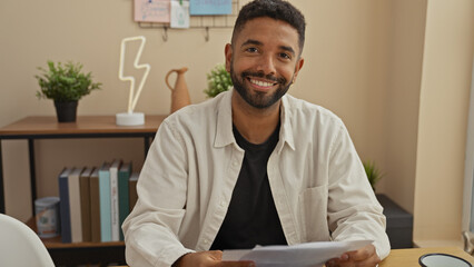 Wall Mural - A young bearded man smiling at home while holding a document, sitting at a wooden desk with decorative plants.