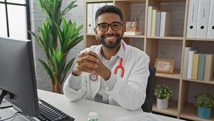 Wall Mural - A cheerful young black man in a white coat with a stethoscope sits at a desk in a modern clinic office