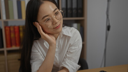 Canvas Print - Chinese woman in office wearing glasses, sitting at desk with hand on cheek, resting in workplace with bookshelves in background.