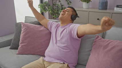 A joyful hispanic man relaxes with headphones on a cozy sofa in a modern apartment.