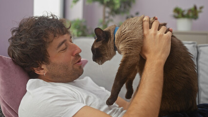 Wall Mural - A young man affectionately pets his siamese cat in a cozy living room, creating an image of comfortable domestic life.