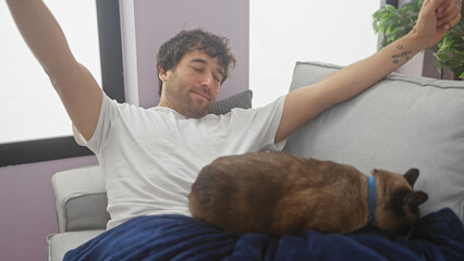 Hispanic man stretching with eyes closed and a cat sleeping beside him on a couch in a cozy apartment living room