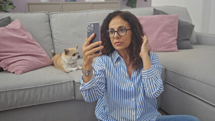 A middle-aged woman checks her smartphone with a chihuahua beside her on the living room sofa at home.