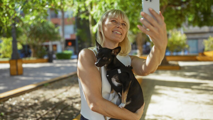 Woman taking selfie in urban park holding chihuahua, with greenery and buildings in the background under bright sunlight.