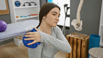 A young hispanic woman applies a cold compress while sitting in a rehab clinic's treatment room.