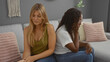 © Krakenimages.com - Women sitting back to back on a sofa in a living room, showing emotions of disagreement and contemplation, with pillows and wall decoration in the background