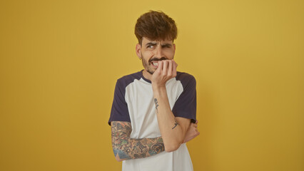 Poster - A worried young man with tattoos stands against a yellow background, biting nails and looking nervous.