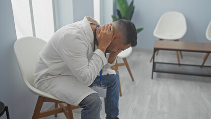 Canvas Print - A stressed hispanic man in a white coat holding his head in a hospital waiting room