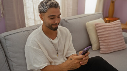 Sticker - Young arab man with a beard sitting on a couch at home, using a mobile phone in a cozy living room with pillows and natural light.