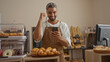 © Krakenimages.com - Young man with beard in bakery wearing apron looking at smartphone celebrating success surrounded by bread and pastries