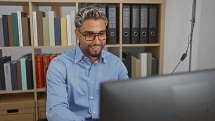 Wall Mural - Young arab man working in an office indoors smiling while sitting at his desk with bookshelves behind him