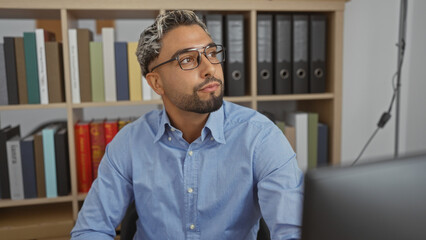 Sticker - Young, arab, muslim man with a beard sitting in an office workspace indoors, surrounded by shelves filled with books and folders