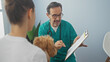 © Krakenimages.com - A middle-aged hispanic man in scrubs consults with a female client holding a poodle inside a veterinary clinic.