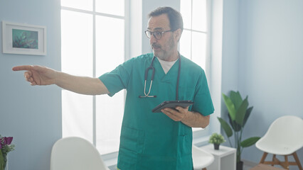 Poster - Hispanic doctor pointing in a bright clinic room while holding a tablet