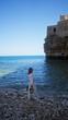 © Krakenimages.com - A young hispanic woman enjoying the beach at polignano a mare, puglia, italy, standing on the rocky shore with clear blue water and a historic stone building in the background.