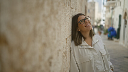 Poster - Beautiful young hispanic woman smiling in the charming streets of polignano a mare, puglia, italy, leaning against a stone wall on a sunny day.