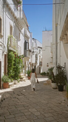 Poster - A young hispanic woman walks through the charming, white streets of the old town in locorotondo, puglia, italy, surrounded by beautiful, historic european architecture on a sunny day.