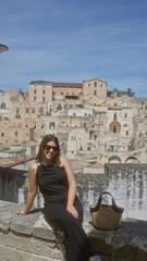 Poster - A beautiful young hispanic woman sits at a viewpoint in the old town of matera, italy, with a charming backdrop of historic buildings.