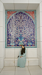 A young adult hispanic woman in traditional islamic clothing sitting against a beautifully patterned mosque wall in abu dhabi.