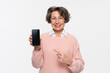 © InsideCreativeHouse - Happy smiling caucasian senior mature woman in glasses casual clothes showing blank mockup smartphone screen using phone while looking at the camera isolated over white background.