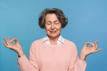 Beautiful calm elderly old woman close eyes smiling feels good folded fingers mudra gesture makes yoga exercise studio head shot isolated on blue background. No stress healthy female retiree concept