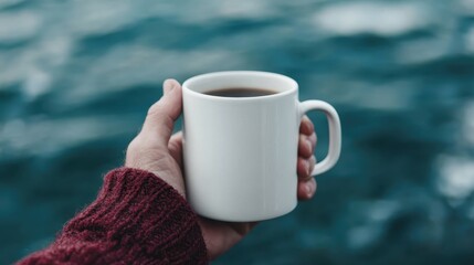 A person in a red sweater holding a white coffee mug with an expansive ocean in the background, capturing a tranquil and refreshing moment near the sea.