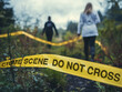 © zphoto83 - Two people are seen walking away from a crime scene marked with yellow tape in a secluded forest, with dense foliage and a cloudy sky creating a somber atmosphere