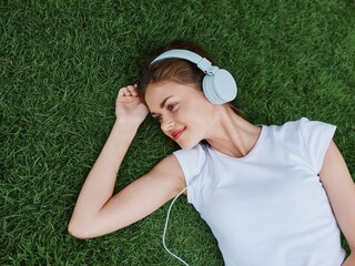 Wall Mural - Woman listening to music with headphones in a white T-shirt lying on the green grass of the lawn in the park, meditating with her eyes closed